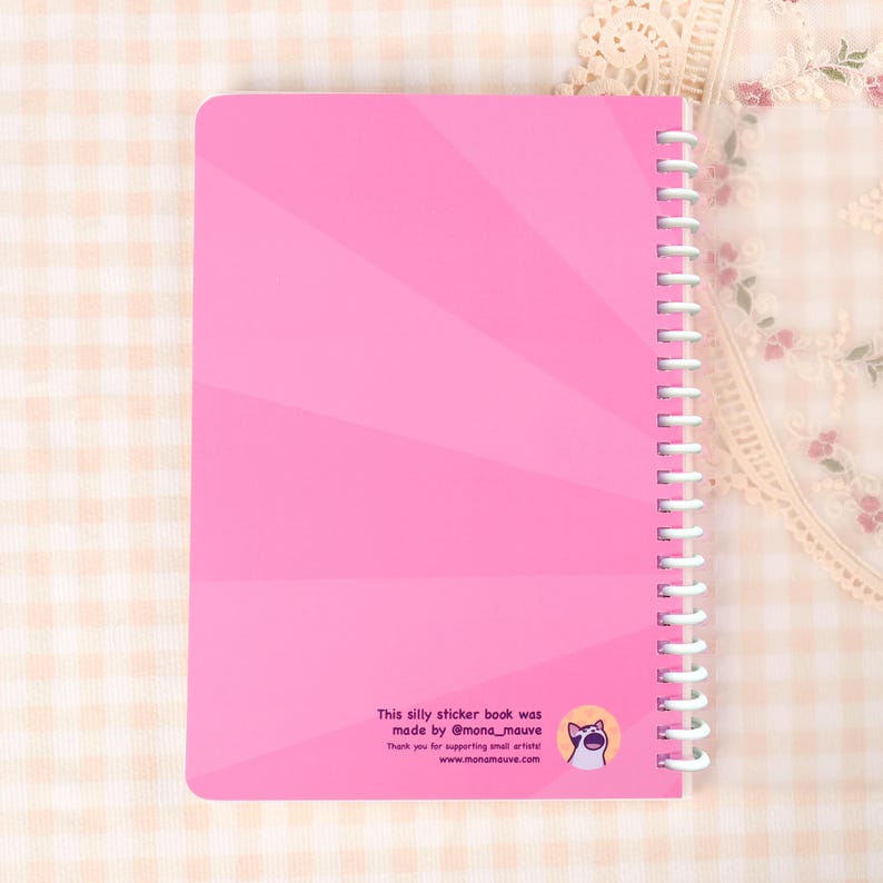 May include: A pink spiral-bound notebook with a radiating design on the cover. The text "This silly sticker book was made by @mona_mauve" is printed at the bottom, along with a cat illustration and website address.