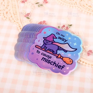 K&ouml;nnte beinhalten: Ein Stapel schimmernder Aufkleber in Wolkenform. Jeder Aufkleber zeigt eine lila Katze mit Hexenhut und Umhang, die auf einem Besen reitet. Der Text lautet "On my way to cause mischief".