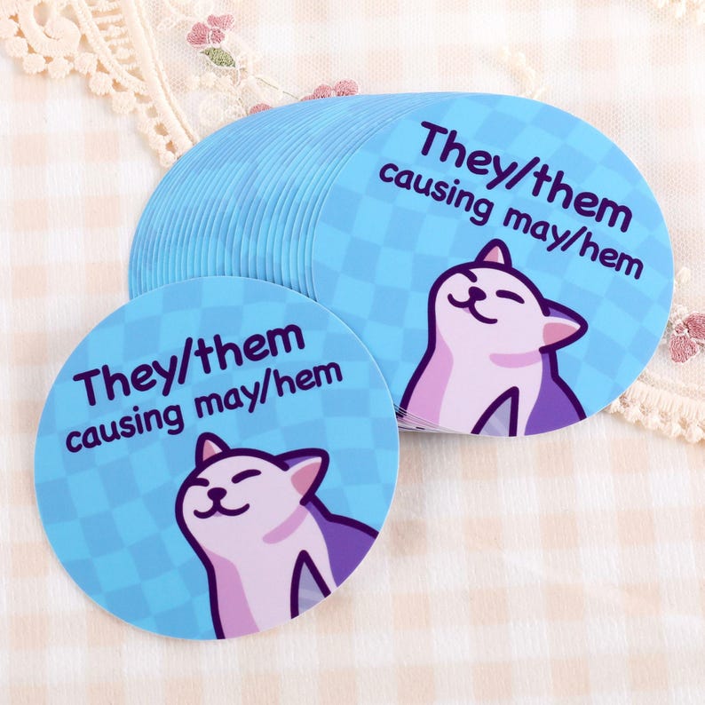 Op de afbeelding: Een stapel lichtblauwe, ronde stickers met een cartoon kat illustratie. De stickers hebben de tekst "They/them causing may/hem" in paars en wit. De achtergrond van de stickers is een lichtblauw geruit patroon.