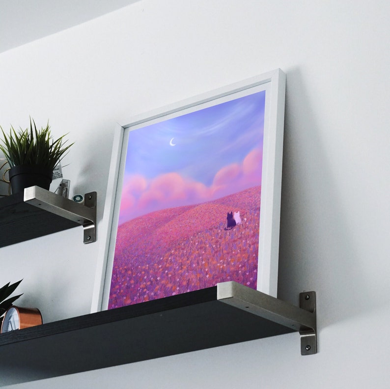 May include: A white framed print of a painting depicting a pink and purple landscape with two black and white cats sitting in a field of flowers under a crescent moon.