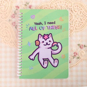 May include: A spiral-bound notebook with a green cover featuring a cartoon cat holding a rainbow and the text "Yeah, I need ALL OF THESE!". The cat has a star on its head and a smiley face on its ear. The background has stars, moons, and other graphics.