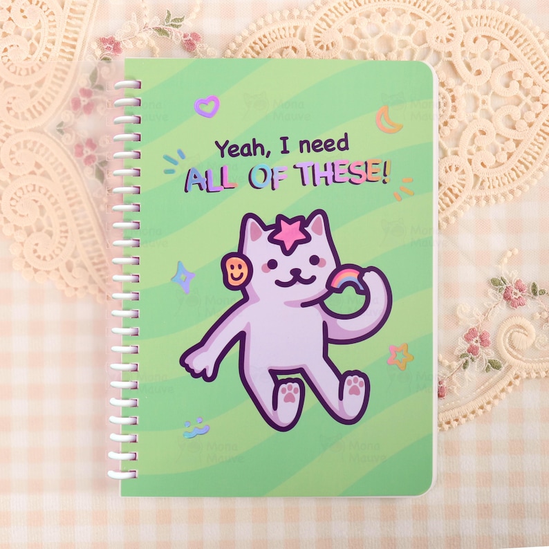 May include: A spiral-bound notebook with a green cover featuring a cartoon cat holding a rainbow and the text "Yeah, I need ALL OF THESE!". The cat has a star on its head and a smiley face on its ear. The background has stars, moons, and other graphics.