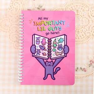 May include: Pink spiral-bound notebook with the text "All my IMPORTANT LIL GUYS go here!!" and a cartoon cat holding a book. The book's pages display various cartoon illustrations. The notebook has a white spiral binding.