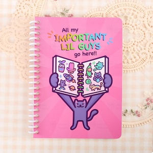 Könnte beinhalten: Ein pinkfarbenes Spiralnotizbuch mit einer lila Katzenillustration, die ein offenes Buch voller bunter Aufkleber hält. Der Text lautet: All my IMPORTANT LIL GUYS go here!! Der Einband hat ein strahlenförmiges Muster in verschiedenen Rosatönen.