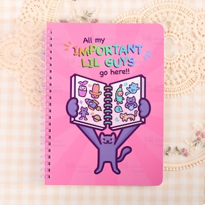 Puede incluir: Cuaderno rosa con espiral con el texto "All my IMPORTANT LIL GUYS go here!!" y un gato de dibujos animados sosteniendo un libro abierto. Las páginas del libro presentan varias ilustraciones de dibujos animados. El cuaderno tiene una cubierta rosa y una encuadernación en espiral blanca.