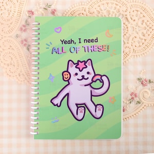 Puede incluir: Un cuaderno con espiral con una cubierta verde que presenta un gato de dibujos animados sosteniendo un arcoíris y el texto "Yeah, I need ALL OF THESE!". El gato tiene una estrella en la cabeza y una carita sonriente en la oreja. Estrellas decorativas, lunas y otras formas rodean al gato.