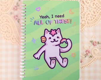 以下が含まれることがあります： 緑色の表紙に、虹を持った漫画の猫と「Yeah, I need ALL OF THESE!」という文字が描かれたスパイラル綴じノート。 猫の頭には星、耳にはスマイリーフェイスがあります。 装飾的な星、月、その他の形が猫を囲んでいます。