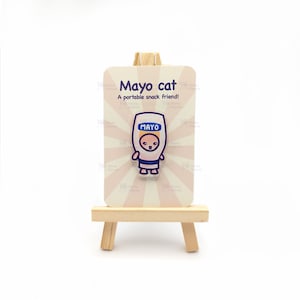 Może przedstawiać: Biała kartka z różowymi paskami i tekstem "Mayo cat" oraz "A portable snack friend!"  Na kartce znajduje się kreskówkowy kot w kostiumie słoika z majonezem. Kartka stoi na małym drewnianym sztalugach.