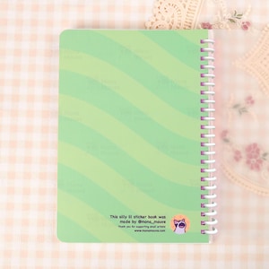May include: A spiral-bound sticker book with a light green cover featuring diagonal stripes. The text "This silly lil sticker book was made by @mona_mauve" is printed on the bottom. A cartoon cat logo is also visible.