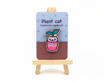 May include: A wooden pin featuring a white cat with a green plant growing out of its head. The cat is in a pink pot with a blue stripe. The pin is on a brown wooden background with the text "Plant cat Eco-friendly wooden pin".