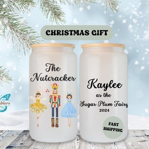 Puede incluir: Dos vasos de vidrio esmerilado con tapas y pajitas de bambú. El vaso de la izquierda tiene un gráfico de una bailarina, el Cascanueces y otra bailarina. El texto "The Nutcracker" está impreso en el vaso. El vaso de la derecha tiene el texto "Kaylee as the Sugar Plum Fairy 2024".