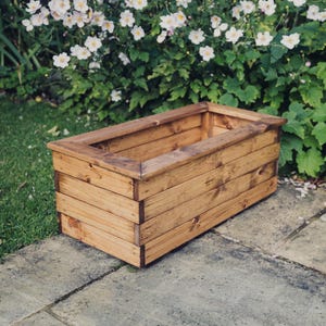 May include: A rectangular wooden planter box with a natural brown stain finish. The planter is made of multiple wooden planks and has a slightly raised lip around the top.