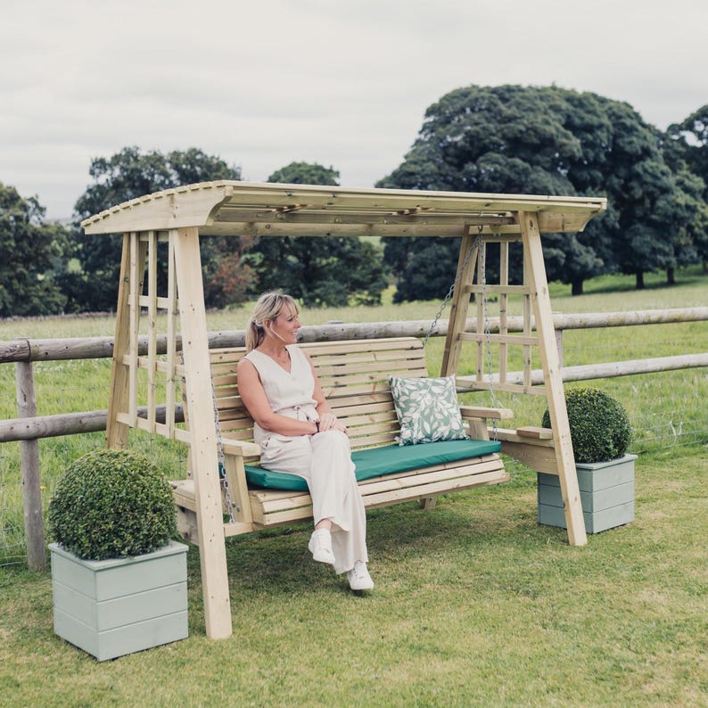 May include: A wooden garden swing bench with a canopy roof, featuring a slatted backrest, a green seat cushion, and a patterned throw pillow. The structure is supported by two side frames, each flanked by a square planter containing a round shrub.
