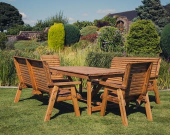 May include: A wooden outdoor dining set with a rectangular table and six chairs. The chairs have slatted backs and are arranged around the table. The set is made of natural wood and has a rustic look. The set is on a green lawn with a lush garden in the background.