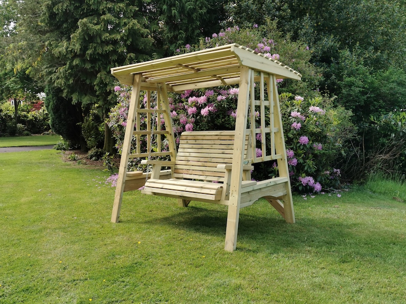 May include: A light-coloured wooden garden swing bench featuring a slatted seat and backrest, suspended by chains from a sturdy frame with a slatted roof canopy and integrated side armrests, positioned on a green lawn in an outdoor setting.