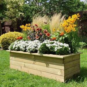 May include: A wooden rectangular planter box overflowing with colourful flowers and foliage. The flowers include white, yellow, orange, and red blooms. The planter is made of light-coloured wood and is set on a green lawn.