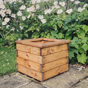 May include: A square wooden planter box with a natural wood finish. The planter is made of multiple planks of wood and has a slightly distressed look.