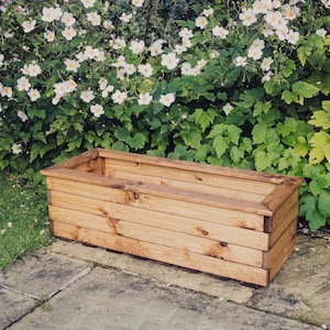 May include: A rectangular wooden planter box with a natural brown finish. The planter is made of multiple horizontal planks of wood and has a slightly raised lip around the top edge.