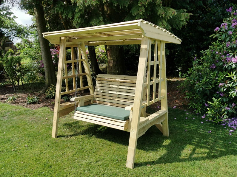 May include: A light-coloured wooden garden swing with a slatted bench seat, a matching overhead canopy roof, and lattice-style side panels. The bench features a solid olive green rectangular cushion and is suspended by metal chains from the frame.