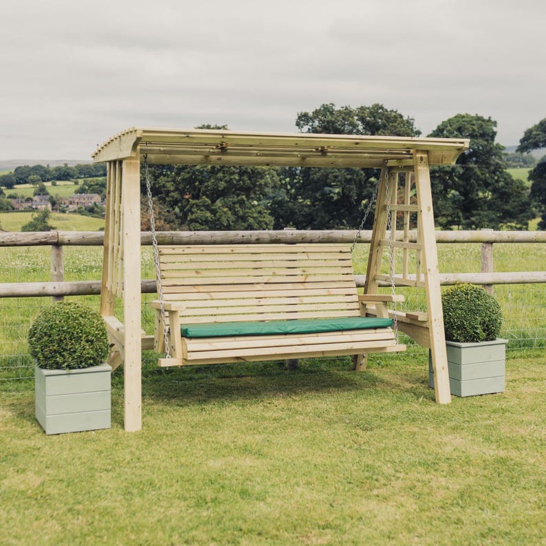 May include: A wooden garden swing bench with a slatted back and seat, suspended by metal chains from a sturdy frame with a roof. The bench features a long, rectangular green cushion. Two light green square planters sit on either side.
