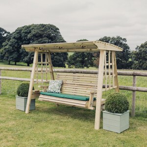May include: A wooden garden swing bench with a canopy roof, featuring a slatted back and seat. The bench includes a long dark green cushion and a single square throw pillow with a white and green leaf pattern, suspended by metal chains.