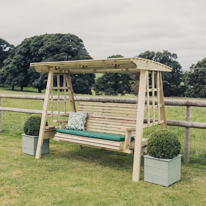 May include: A wooden garden swing bench with a canopy roof, featuring a slatted back and seat. The bench includes a long dark green cushion and a single square throw pillow with a white and green leaf pattern, suspended by metal chains.