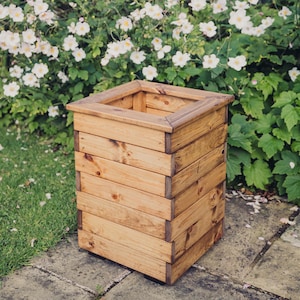 May include: A square wooden planter box with a natural wood finish. The planter is made from multiple horizontal planks of wood.