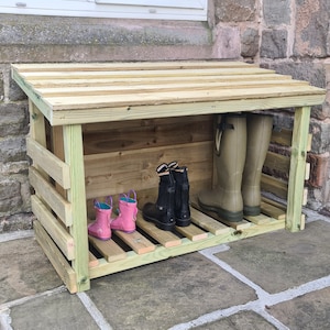 May include: A wooden boot rack with a sloping roof, offering protection for footwear. It has several shelves for storing boots and shoes, with a pair of green rubber boots and two pairs of pink and black children's boots visible.