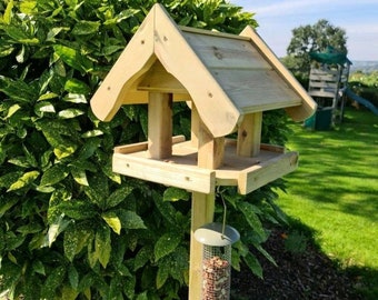 May include: A wooden bird feeder with a peaked roof and two feeding platforms. The feeder is mounted on a pole and has a metal seed dispenser hanging below.