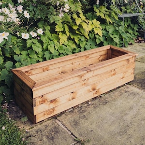 May include: A rectangular wooden planter box made from light-coloured wood. The box is constructed with horizontal planks and has a simple, open design. It sits on a stone surface, with greenery and flowers in the background.