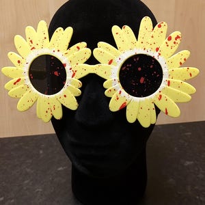 May include: Yellow daisy-shaped sunglasses with red splatter paint. The sunglasses are on a black mannequin head.