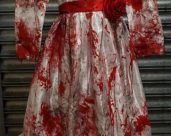 Könnte beinhalten: Weißes Kleid mit langen Ärmeln und rotem Schärpe, bespritzt mit roter Farbe. Eine große rote Rose ist an der Taille befestigt. Das Kleid hat einen Rüschensaum und wird auf einer Schaufensterpuppe präsentiert, für Halloween.