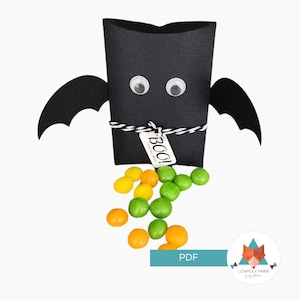 Peut inclure: Un porte-bonbons en papier noir en forme de chauve-souris avec des yeux globuleux et des ailes attachées. Le support est noué avec une ficelle noire et blanche et une étiquette qui dit "BOO!". Des bonbons colorés s'échappent du bas.