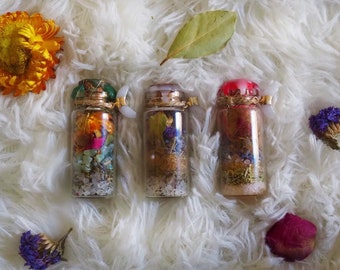 May include: Three small glass bottles filled with dried herbs, flowers, and crystals. The bottles have gold lids and are arranged on a white fluffy surface.