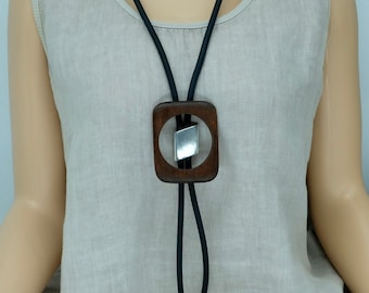 May include: A modern bolo tie featuring a dark wood square pendant with a silver accent. The pendant is suspended from a black cord. The tie is worn over a neutral-colored sleeveless top.