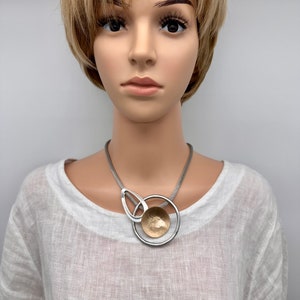 May include: A silver and gold necklace with a large, round pendant. The pendant is made of two circles, one silver and one gold, with a smaller gold circle in the centre. The necklace is worn by a mannequin with short blonde hair.