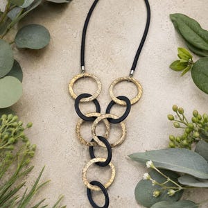 May include: A necklace featuring a black cord and a series of interlocking circular rings. The rings alternate between a textured gold-tone finish and a smooth matte black finish, arranged in a cascading vertical pattern that tapers at the bottom.