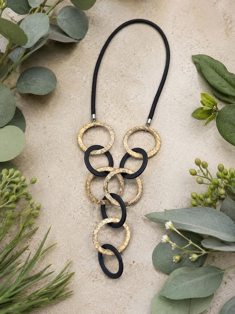 May include: A necklace featuring a black cord and a series of interlocking circular rings. The rings alternate between a textured gold-tone finish and a smooth matte black finish, arranged in a cascading vertical pattern that tapers at the bottom.
