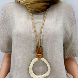 May include: A beige linen shirt with a long necklace featuring a large, off-white circular pendant. The necklace has a brown wooden bead and a smaller amber-coloured bead. A tan scarf is draped around the neck.