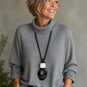 Peut inclure: Un long collier à cordon noir avec une perle rectangulaire blanche et un grand pendentif ovale noir brillant. Le collier est porté sur un pull gris à manches longues avec un col roulé côtelé et des boutons métalliques sur les poignets.
