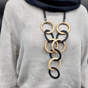 May include: A long necklace featuring a series of interlocking circular rings in alternating matte black and textured gold finishes. The rings are suspended from a thick black cord, creating a cascading geometric pattern that hangs against a light gray linen top.