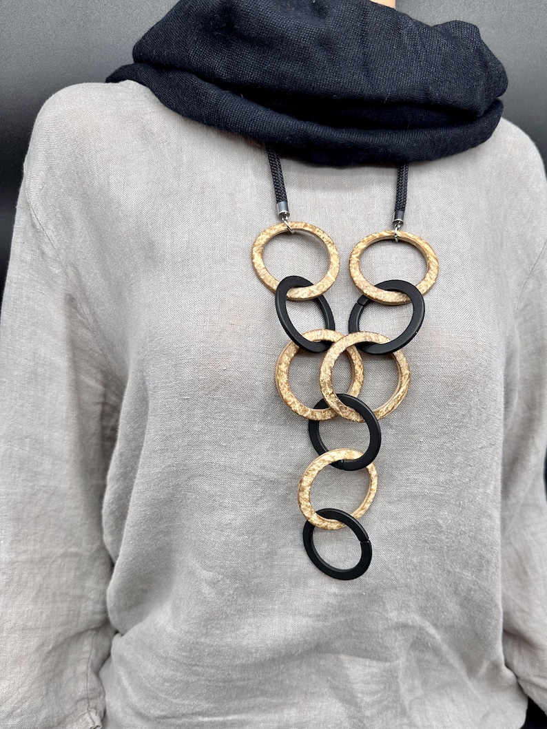 May include: A long necklace featuring a series of interlocking circular rings in alternating matte black and textured gold finishes. The rings are suspended from a thick black cord, creating a cascading geometric pattern that hangs against a light gray linen top.