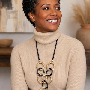 May include: A long necklace featuring a black cord and a series of interlocking circular pendants. The design alternates between textured gold-toned metal rings and smooth matte black rings, arranged in a cascading vertical pattern down the center.