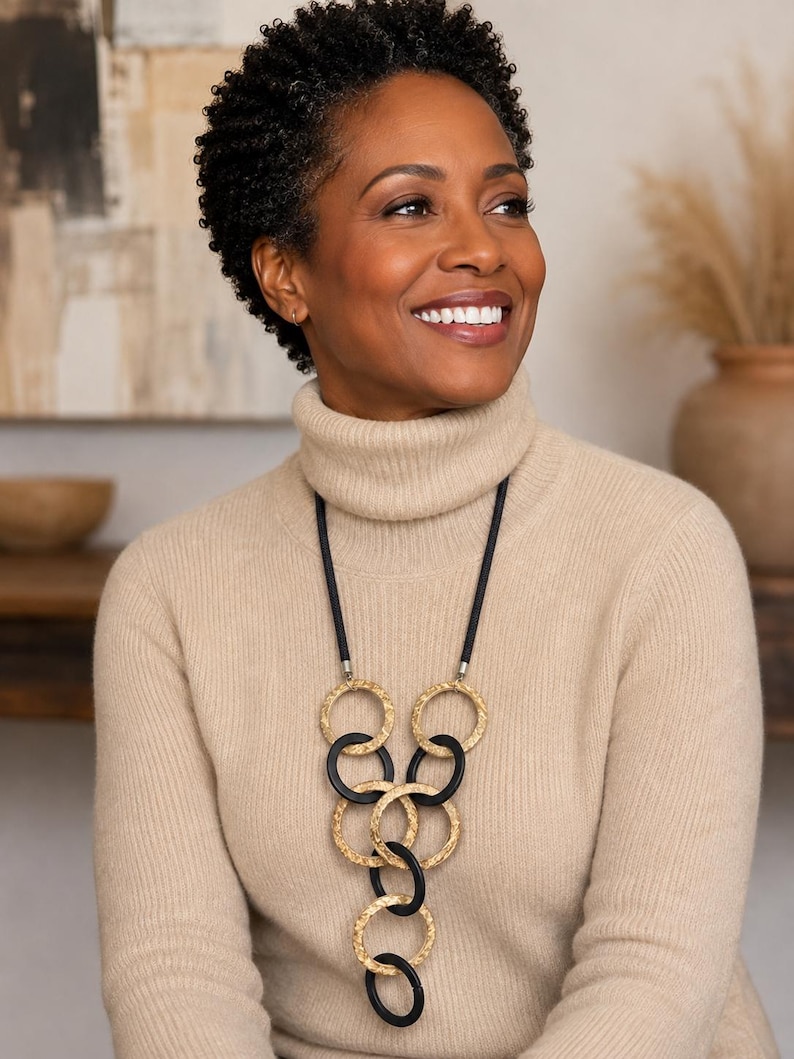 May include: A long necklace featuring a black cord and a series of interlocking circular pendants. The design alternates between textured gold-toned metal rings and smooth matte black rings, arranged in a cascading vertical pattern down the center.