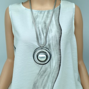 May include: A silver necklace with a large, round pendant featuring concentric circles and a pearl-like centre. The necklace is worn by a model wearing a white top with a black and grey abstract design.