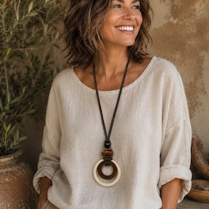 Peut inclure: Un chemisier en lin beige à manches longues avec une encolure ronde et des poignets retroussés, accompagné d'un long collier composé d'un cordon marron foncé, d'une perle noire, d'une perle marron et de deux grands pendentifs circulaires.