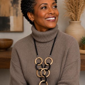 May include: A long necklace featuring a black cord and a series of interlocking circular pendants. The design alternates between textured gold-tone metal rings and smooth matte black rings, arranged in a vertical, cascading geometric pattern.