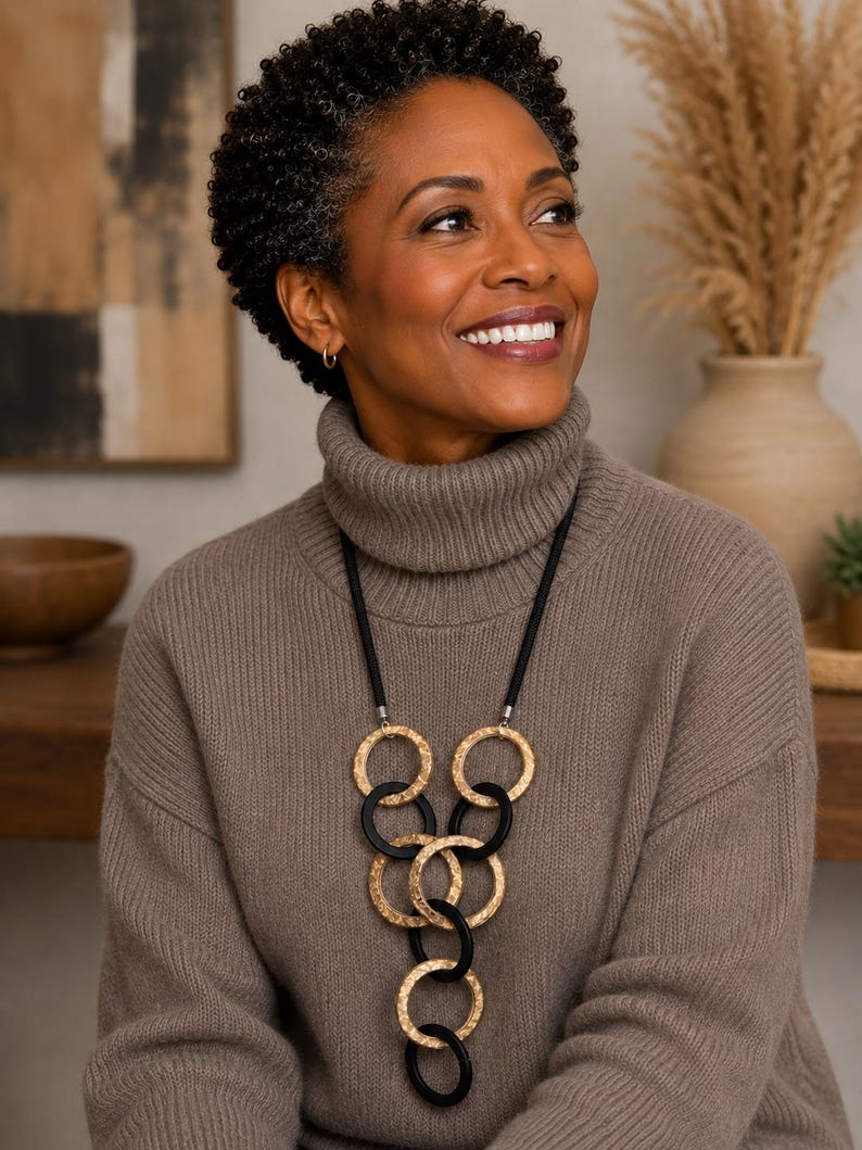 May include: A long necklace featuring a black cord and a series of interlocking circular pendants. The design alternates between textured gold-tone metal rings and smooth matte black rings, arranged in a vertical, cascading geometric pattern.