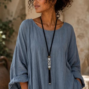 May include: A long necklace featuring a black cord, a central cylindrical pendant with white thread wrapped around a dark metal frame, and two small metal end caps at the bottom of the cord, worn over a loose-fitting blue linen-textured dress.