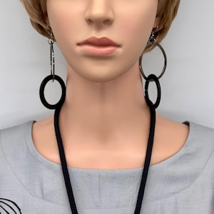 May include: A mannequin wearing a silver and black necklace and earrings set. The earrings feature a large black circle connected to a silver hoop. The necklace has a black rope-like cord with silver accents and large silver circles.